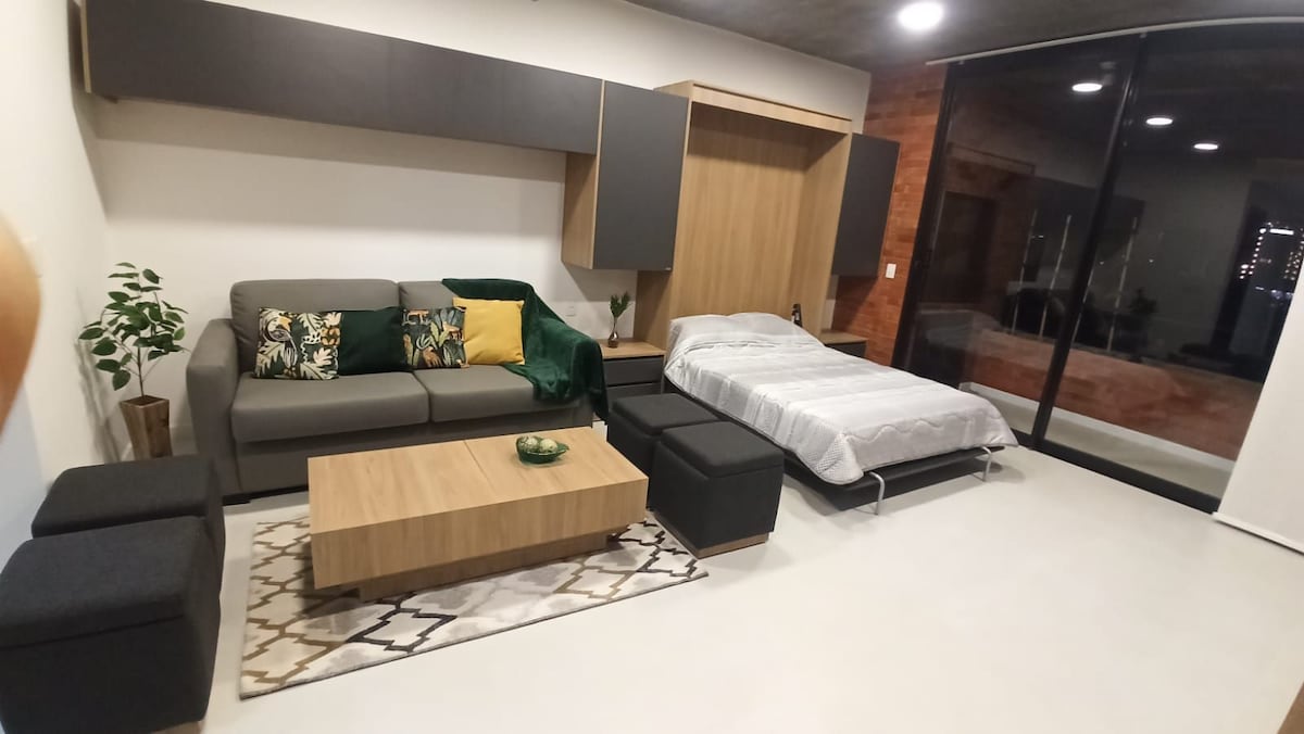 A multifunctional living space features a gray sofa adorned with colorful cushions and a light wooden coffee table. A queen-sized bed is integrated into the cabinetry, with soft bedding neatly arranged. Large glass doors provide views of the outside, allowing natural light to fill the room.