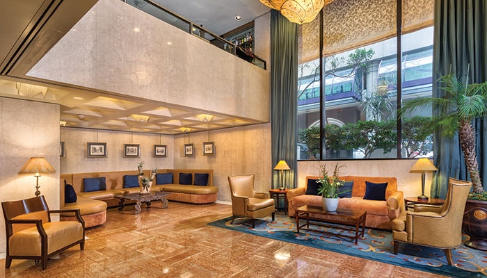 A spacious hotel lobby features elegant marble flooring, with cozy seating areas that include armchairs and a sofa. A variety of plants adds greenery, while large windows allow natural light to brighten the space. Light fixtures hang from the ceiling, contributing to a welcoming ambiance.