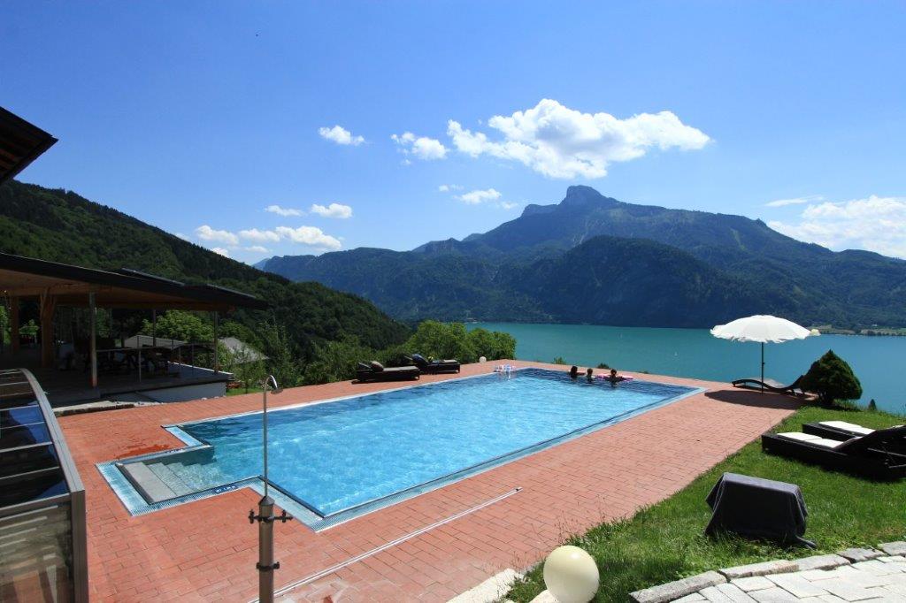 An outdoor heated pool is surrounded by a paved terrace with sun loungers and an umbrella. The scenic view of the lake and mountains highlights the natural beauty of the area, while lush greenery frames the poolside, providing a tranquil atmosphere.