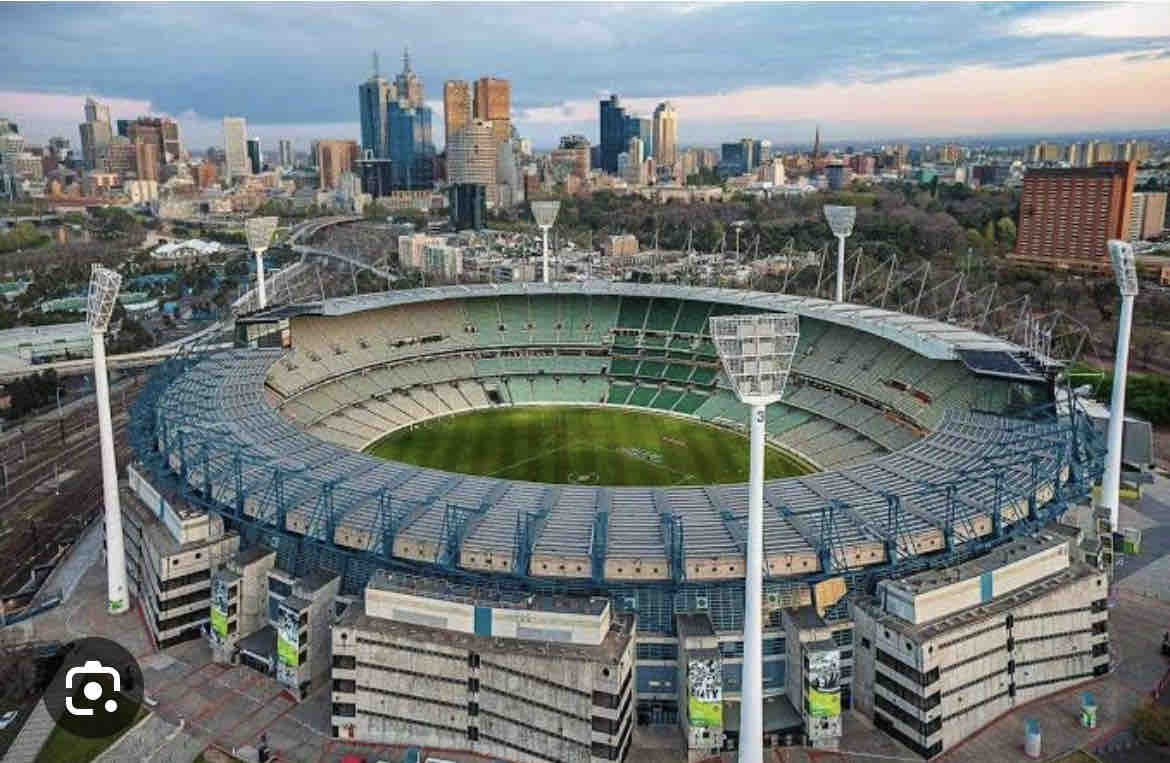 A panoramic view of a large stadium, with its open field visible and seating tiers surrounding the area. The stadium's exterior features modern lighting structures, set against a backdrop of the city skyline showcasing various buildings under a soft gradient sky.