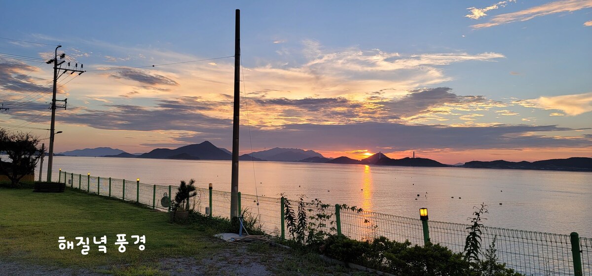 A serene waterfront view is captured, showcasing a sunset over calm waters. Silhouettes of distant islands are visible against a vibrant sky, painted with warm hues. Greenery lines the shore, complementing the tranquil atmosphere.