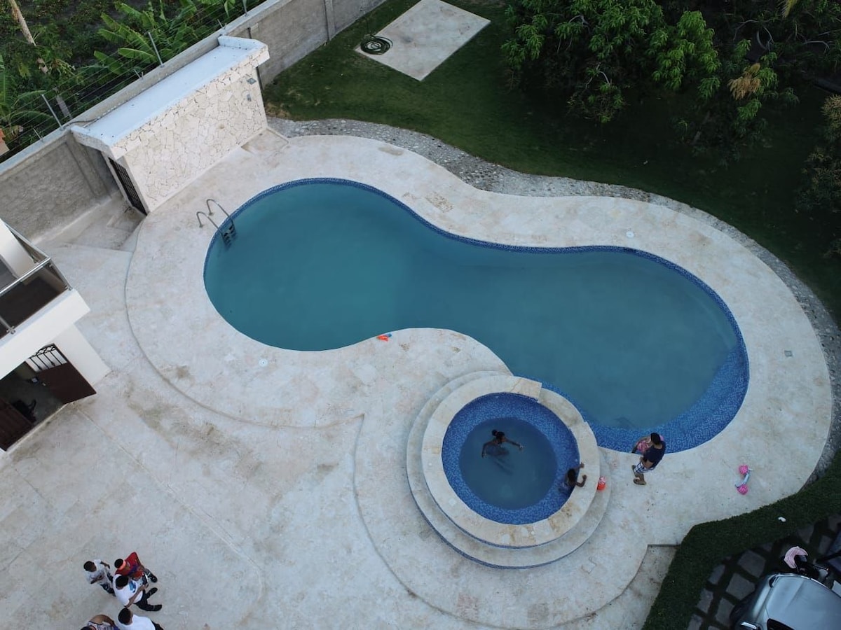An aerial view captures a large, curved swimming pool with a connected jacuzzi, surrounded by a stone patio. Lush greenery borders the area, providing a serene backdrop. A few individuals can be seen enjoying the water, creating a lively yet relaxing environment.