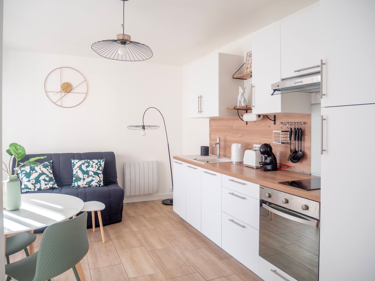 A modern kitchen and living area is depicted, featuring a sleek countertop and built-in appliances. A cozy sofa is arranged near a round dining table, complemented by decorative cushions. Ambient lighting is provided by a unique lamp, enhancing the minimalist style of the space.