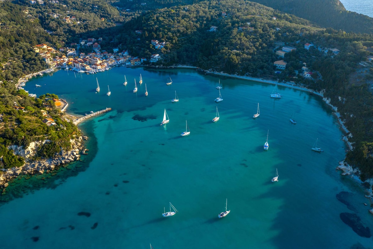 An aerial view captures the calm waters of Lakka port, dotted with sailboats. The coastline is framed by lush greenery and hills, with vibrant residential areas visible in the distance. The blue hues of the sea blend seamlessly with the greenery, creating a serene landscape.