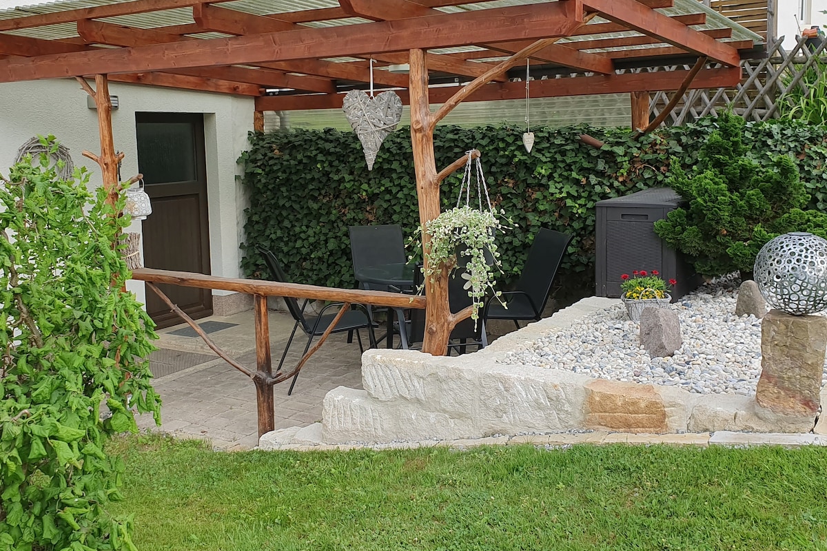 An outdoor terrace is covered by a wooden pergola, featuring four chairs arranged around a table. Lush greenery surrounds the area, alongside a decorative stone feature with pebbles and plants. A storage unit and hanging planters add a touch of charm to the space.