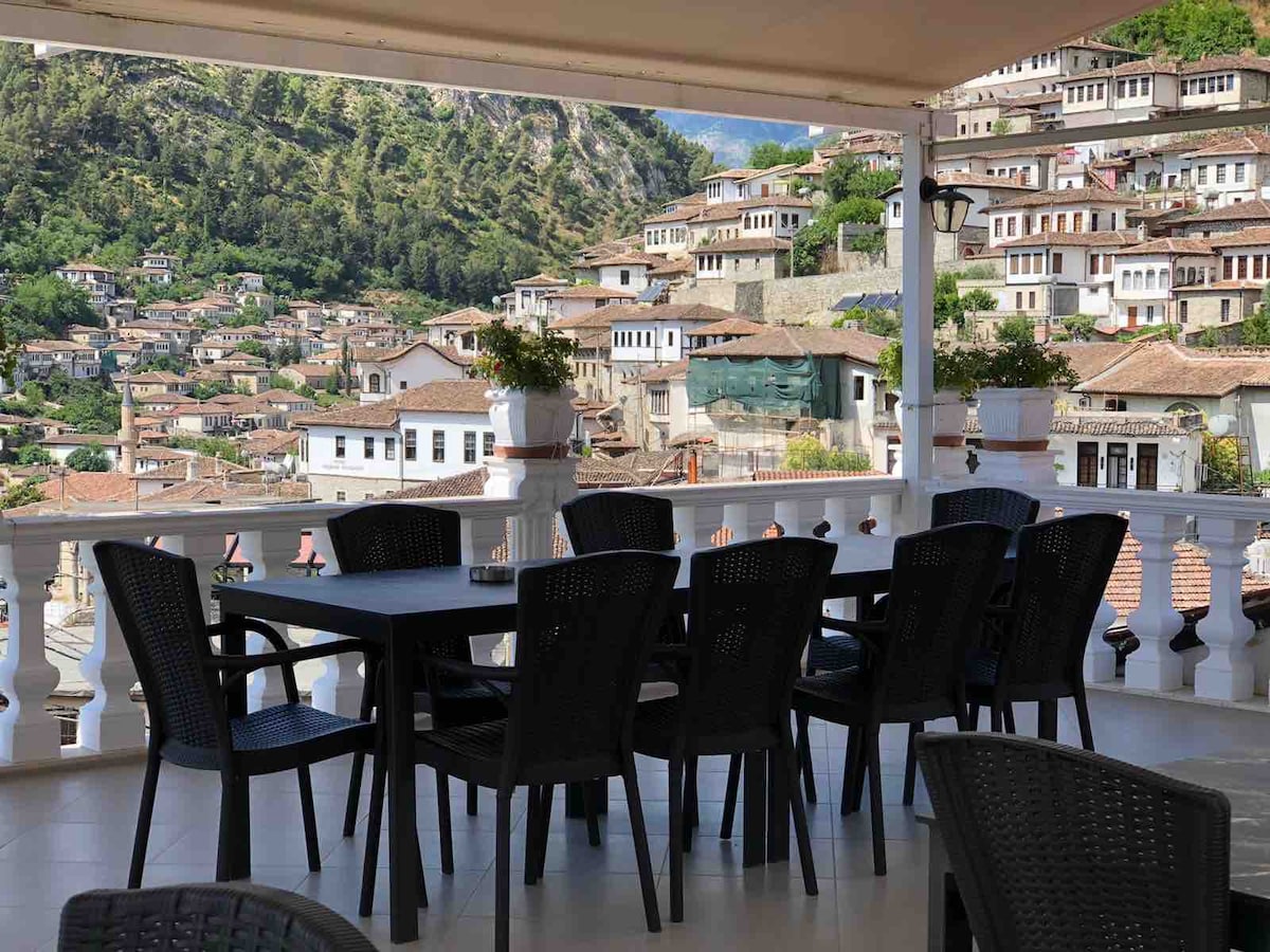 A spacious terrace is furnished with black chairs and tables, offering a scenic view of traditional houses nestled on the hillside. Lush greenery is visible in the distance, showcasing the beauty of the surrounding landscape.