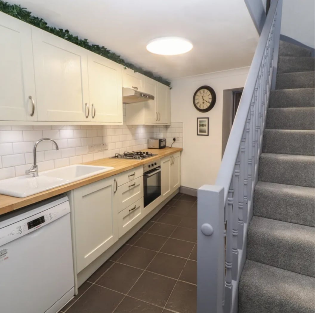 A modern galley kitchen is displayed, featuring solid oak worktops and a ceramic sink. Built-in appliances include an oven, gas hob, and dishwasher. Soft lighting from a ceiling fixture illuminates the space. A staircase is visible in the background, leading to the upper floors.