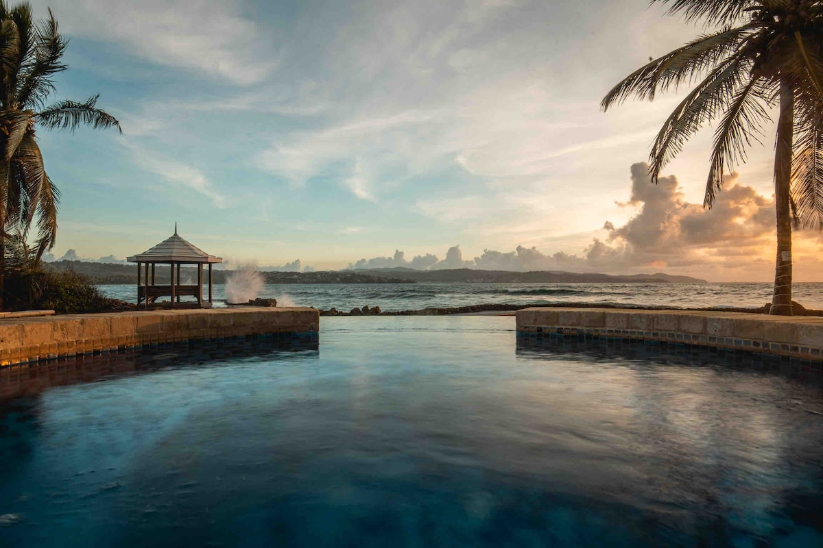 An infinity pool reflects the serene ocean and sky as the sun sets in the background. A gazebo is positioned nearby, surrounded by palm trees. Gentle waves can be seen lapping at the shore, creating a peaceful coastal atmosphere.