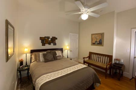 A cozy bedroom is featured with a spacious bed covered in neutral tones. Two bedside lamps provide warm lighting, and a small seating area is present with a wooden bench. A wall-mounted piece of art adds a touch of decor, enhancing the room's inviting ambiance.