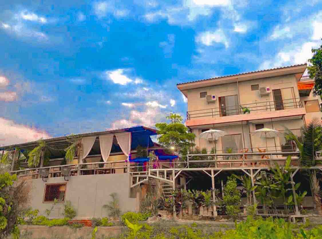 A multi-level structure is visible, featuring wooden balconies adorned with umbrellas. Lush greenery surrounds the area, complementing the spaces. The sky showcases a blend of blue and gray tones, creating a serene backdrop for the building.
