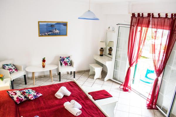 A well-lit room features a spacious bed dressed in red linens. Two chairs with floral patterns are positioned near a white table. A mirror and decorative items adorn a nearby dresser. Large glass doors open to a terrace, with sheer red curtains softly framing the view.