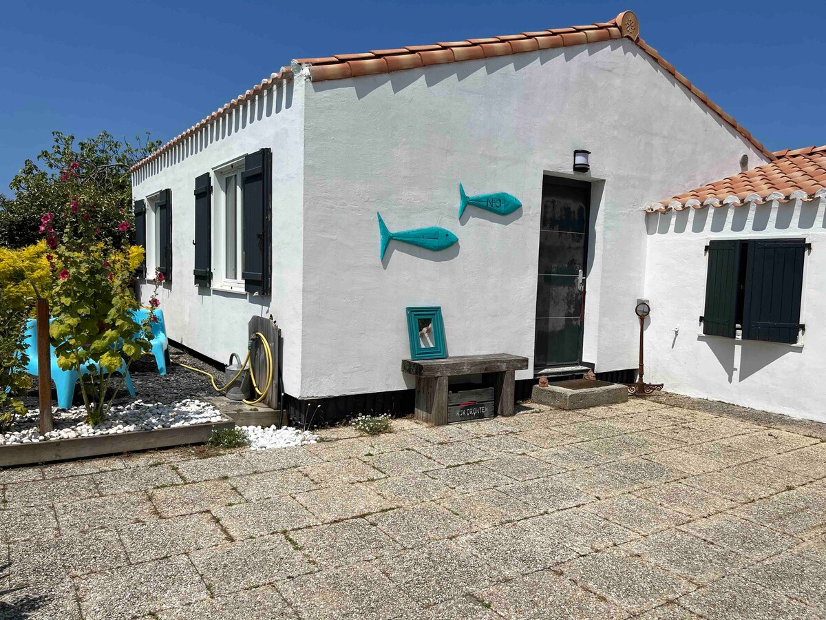 The exterior of the house presents a clean, white facade with decorative blue fish sculptures. A small stoop leads to the entrance, framed by two dark green shutters. The spacious stone-paved area is bordered by vibrant greenery and flowers, enhancing the inviting atmosphere.