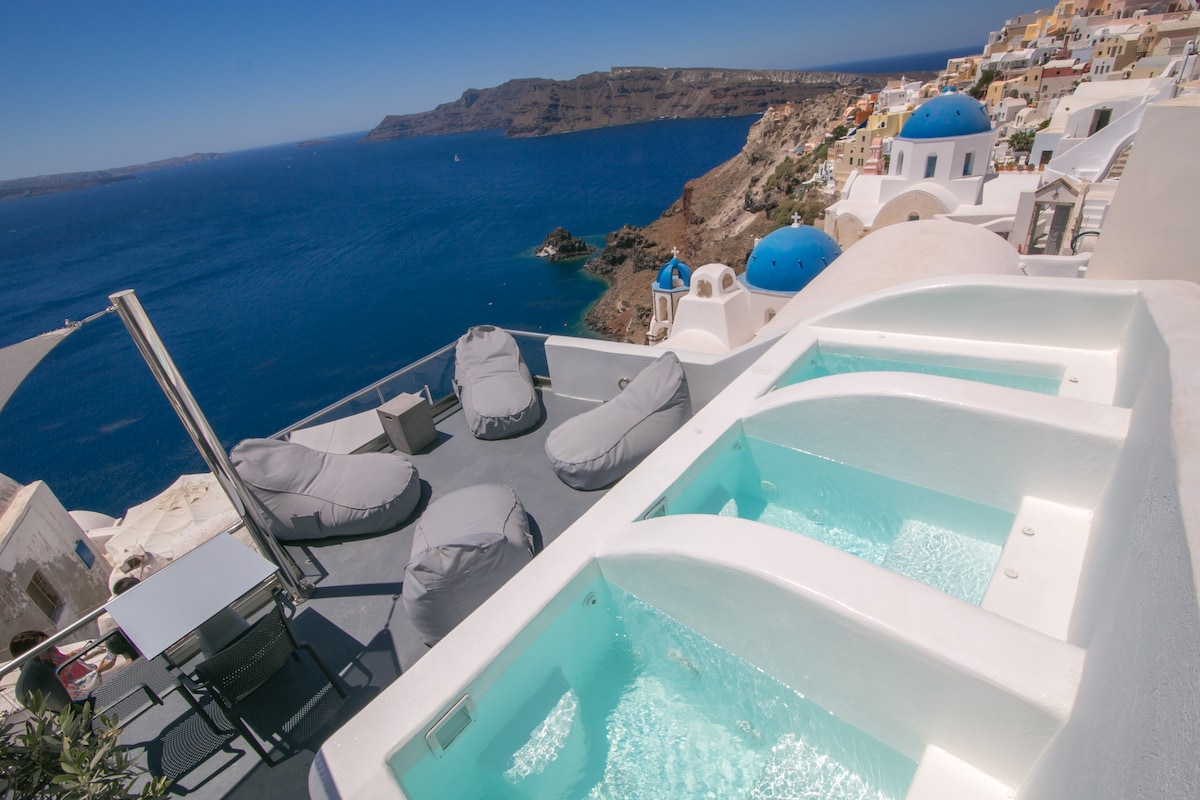 Two private hot tubs are positioned on a spacious terrace, overlooking the Aegean Sea. Soft lounge chairs offer comfortable seating, while clear blue water reflects the sunlight. The landscape features steep cliffs and whitewashed buildings with blue domes, creating a tranquil coastal setting.