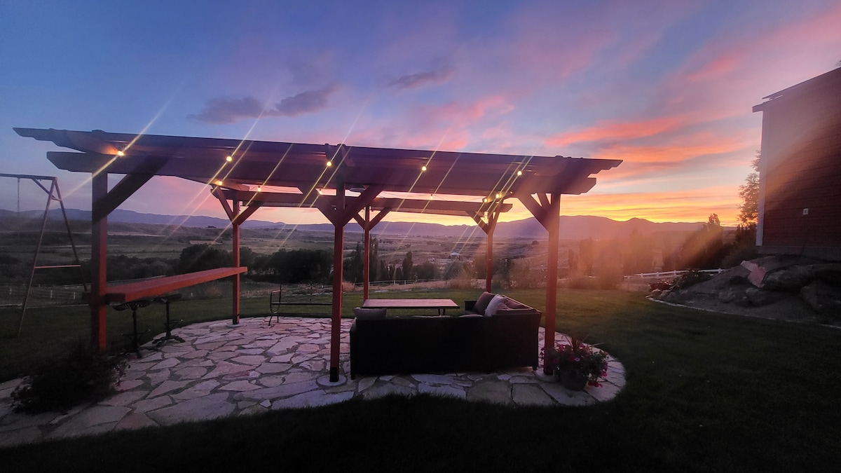 A spacious outdoor patio area features a pergola with soft lighting, ideal for evening gatherings. The view includes rolling hills at sunset, with vibrant colors blending across the sky. A stone pathway leads to comfortable seating, enhancing the serene atmosphere.