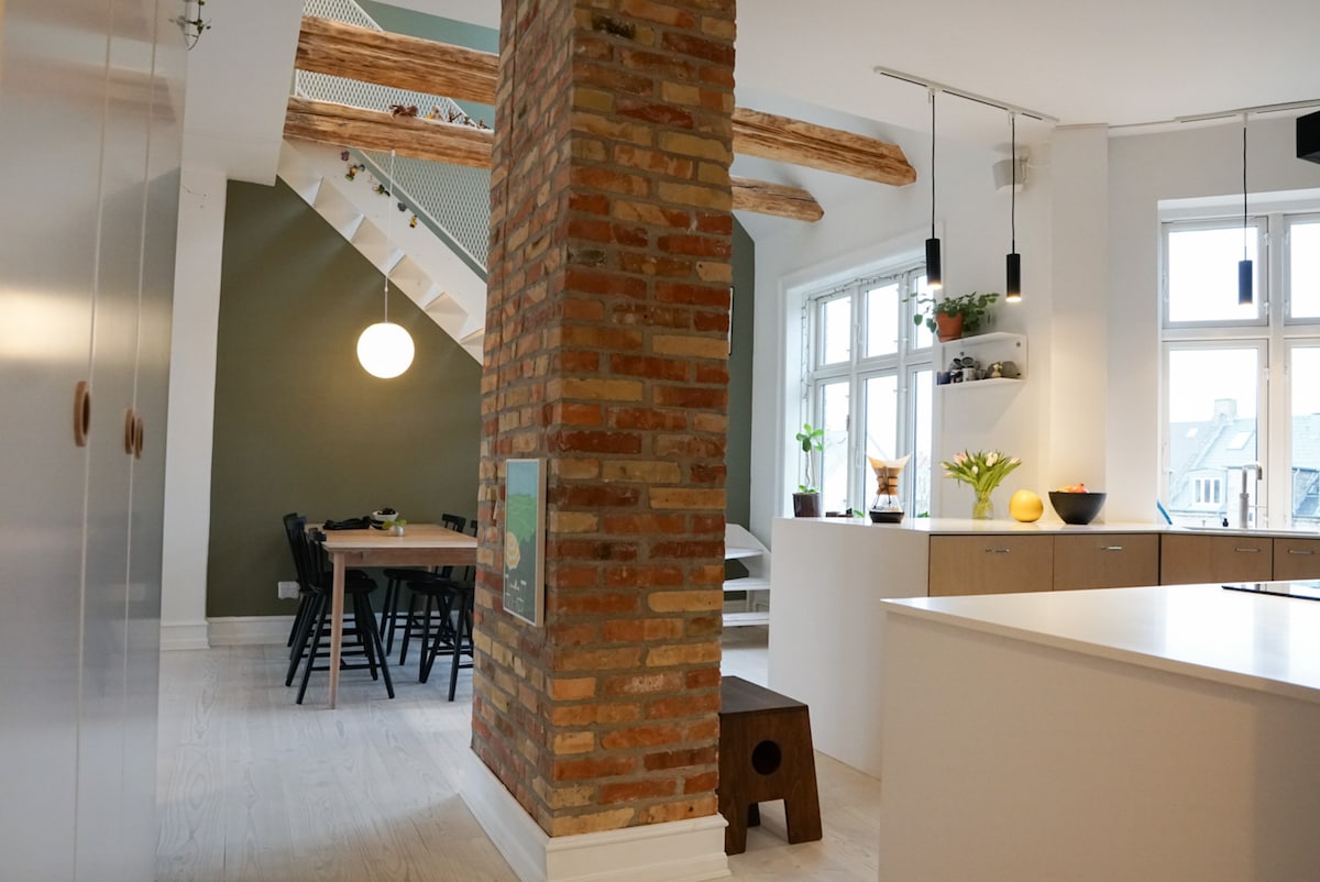 An open kitchen and dining area is showcased, featuring a central island with bar seating. Natural light flows through large windows, highlighting the cozy dining table set for six. Exposed brick and wooden beams add warmth, while modern lighting fixtures enhance the contemporary design.