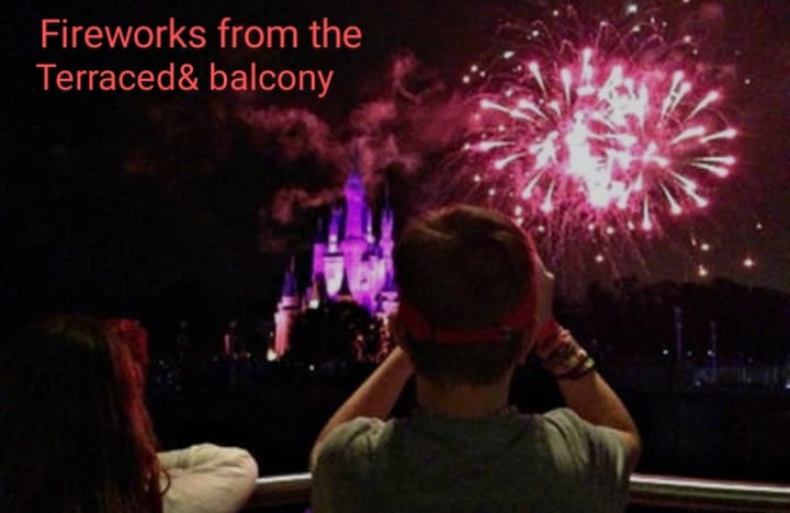 A balcony view captures a vibrant display of fireworks illuminating the night sky. The colorful explosions are set against a distant castle, providing a striking contrast of lights and dark. Two figures are seen here, enjoying the spectacle.
