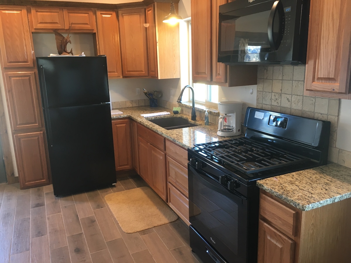 The kitchen showcases modern appliances including a black refrigerator and microwave, alongside a gas stove with an oven. Wooden cabinets provide ample storage, while a granite countertop offers workspace. Natural light enters through a window above the sink, enhancing the space's airy feel.