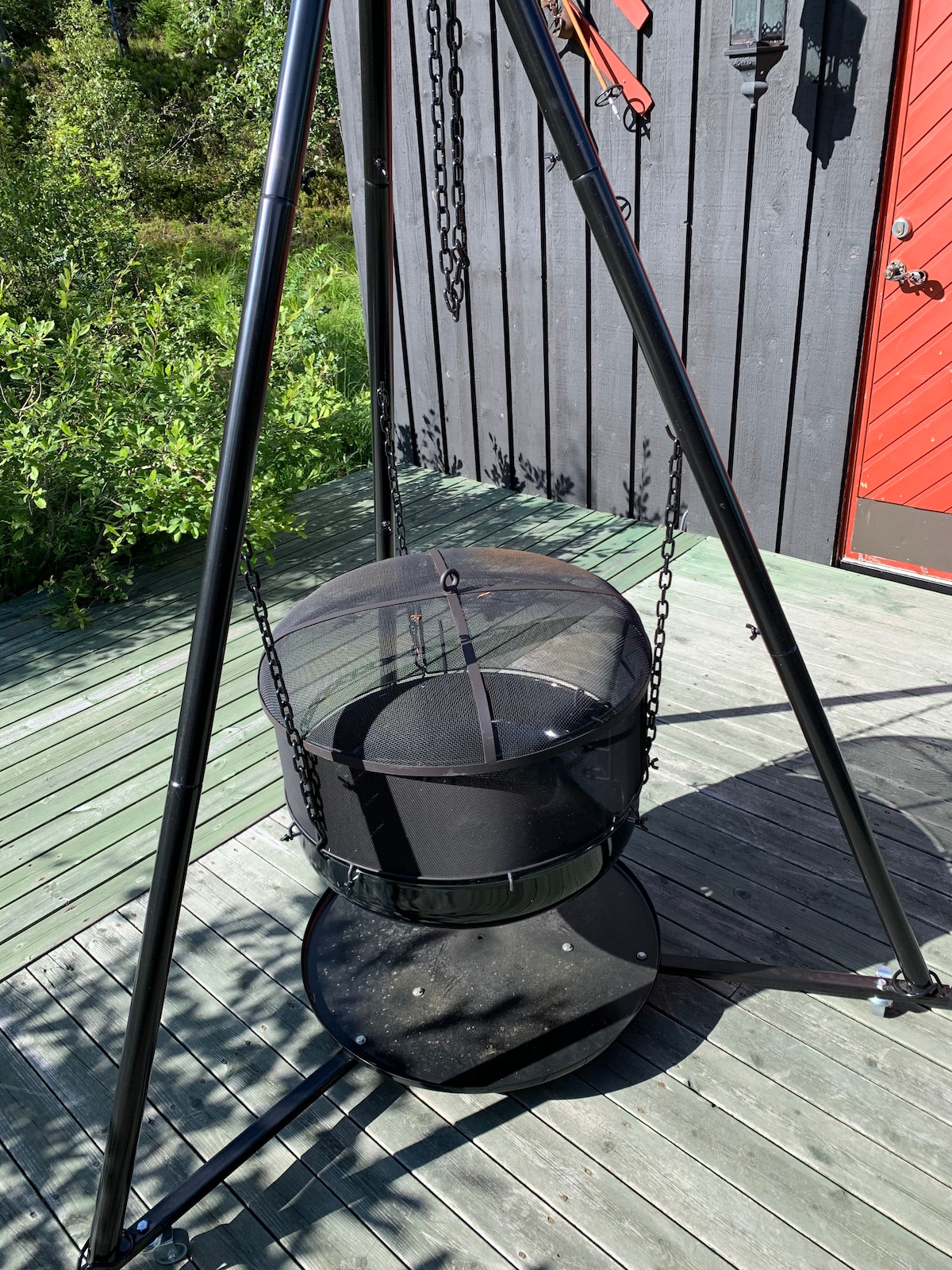 A circular hanging chair is suspended from a sturdy black frame, featuring a soft black mesh cover. The chair rests on a solid base, set against a backdrop of wooden decking and a rustic red door. Green foliage is visible in the surrounding area.