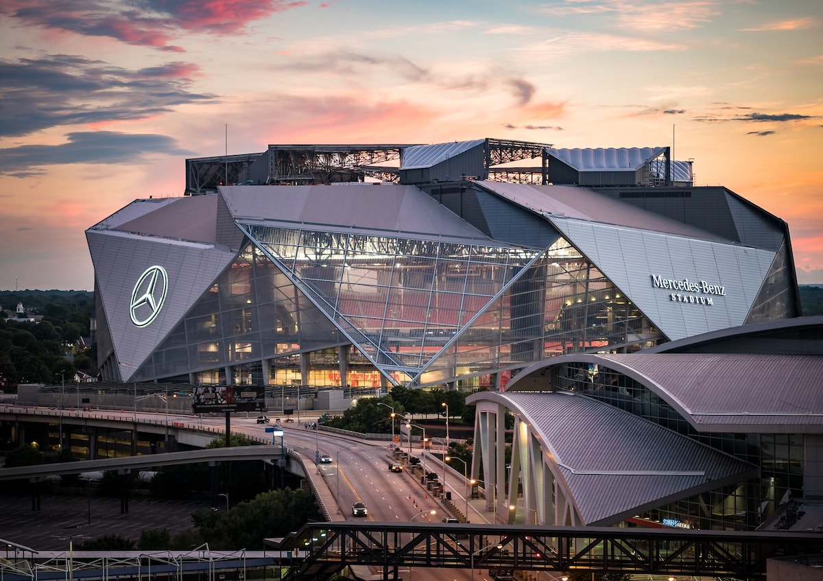 A modern architectural structure, known as Mercedes-Benz Stadium, is depicted at dusk, with dynamic angular designs and large illuminated signages. The building is surrounded by roads and greenery, under a colorful sky as the day transitions to night.