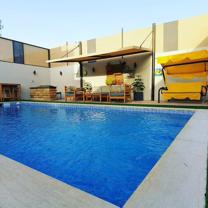 A private heated swimming pool is surrounded by a spacious patio area, featuring lounge chairs and a shaded seating area. Vibrant greenery is visible, alongside a barbecue station. Bright yellow awnings provide shade, contributing to a relaxed outdoor setting, illuminated by natural light.