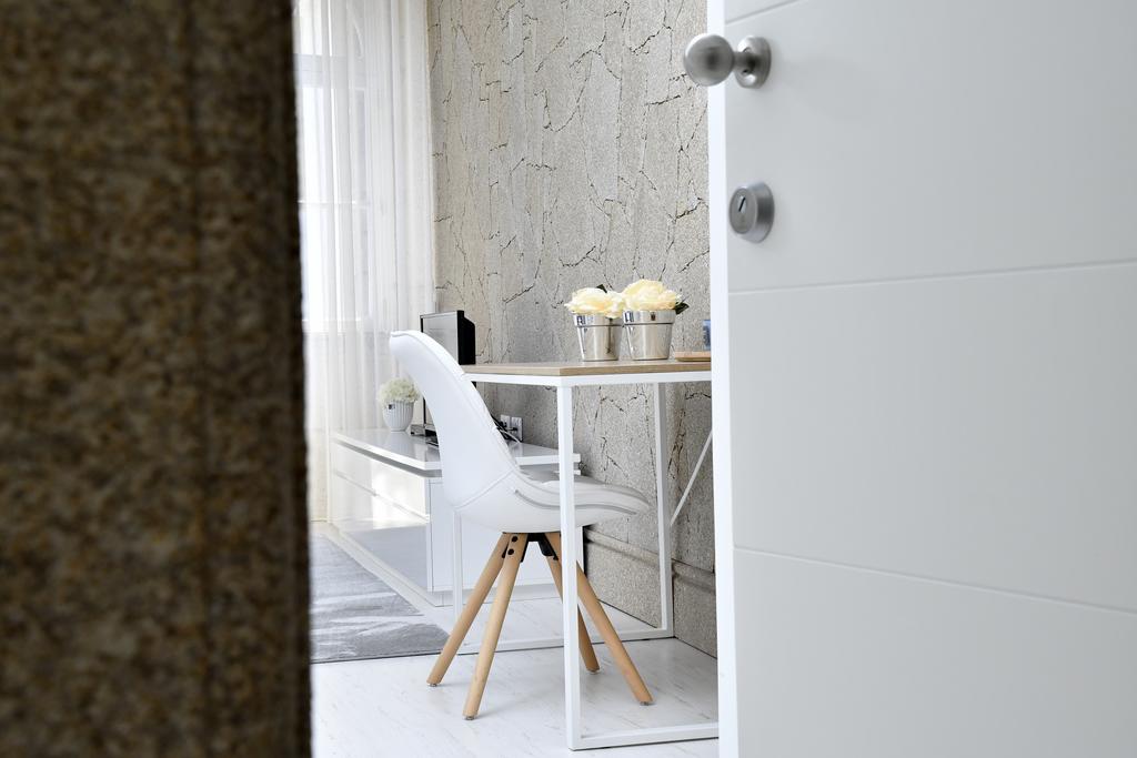 A workspace is visible through a partially open door, featuring a small wooden table with modern white chairs. Two metallic containers are placed atop the table, and a desk is positioned against the wall, offering a minimalist aesthetic with light-colored finishes.
