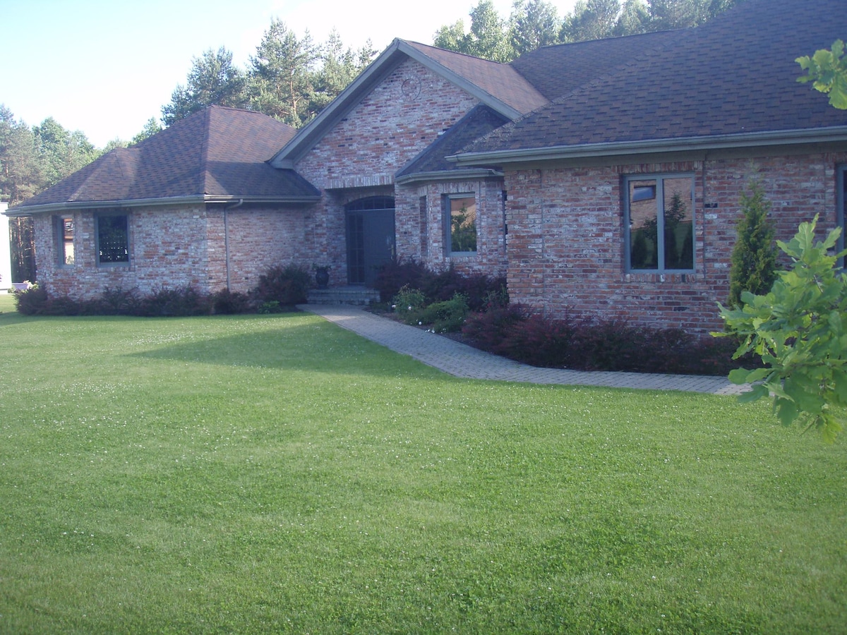 A brick house with a gabled roof is surrounded by lush green grass and trees. The entrance features a walkway made of stone pavers leading up to a front porch, with windows framed by neatly trimmed shrubs.