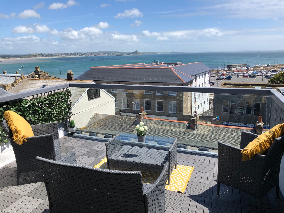 A south-facing balcony features a glass railing and comfortable seating for four, with gray wicker chairs and a small glass table. Two yellow cushions add a pop of color, while views of Mounts Bay and the coastline stretch into the distance.