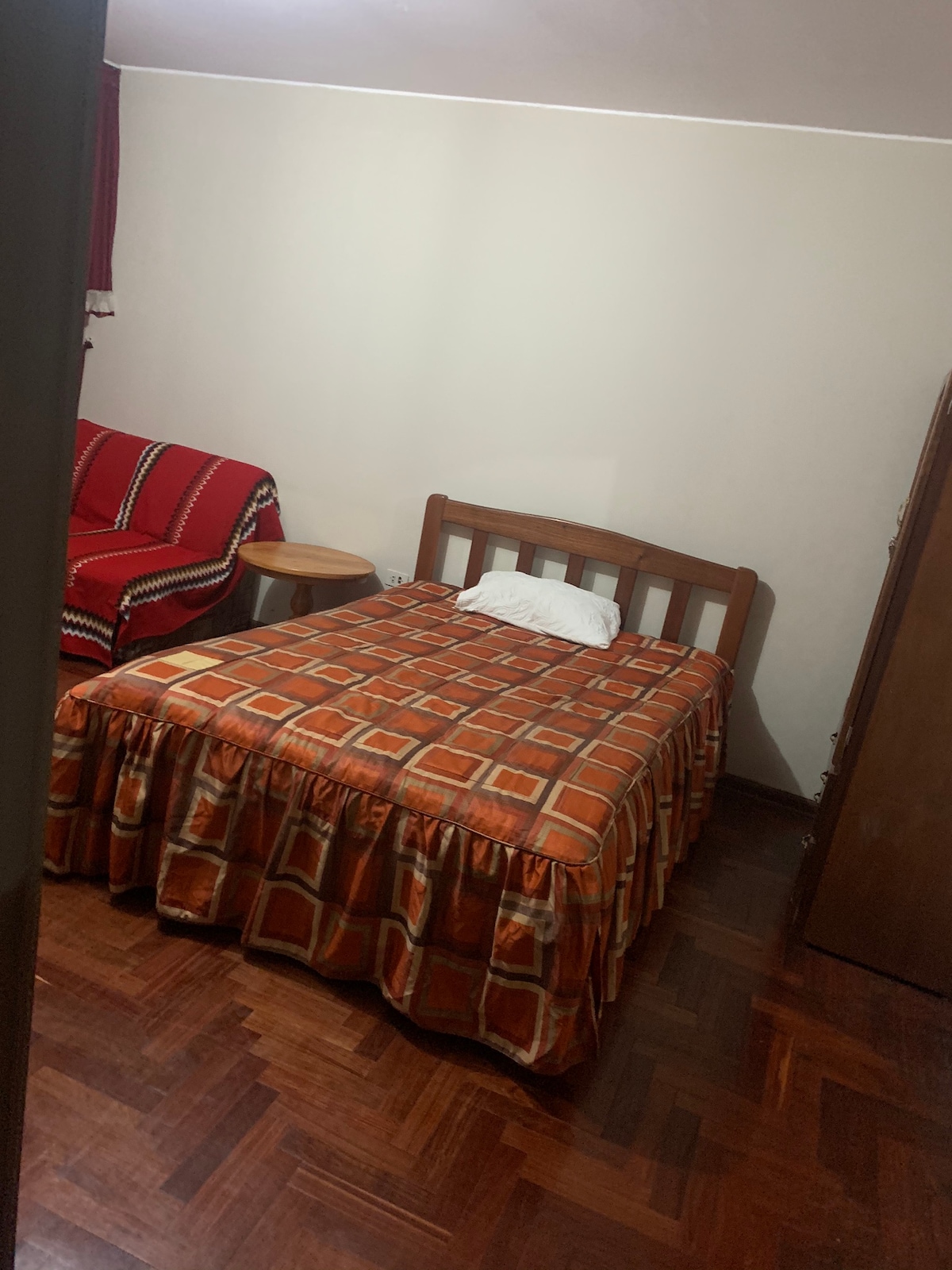 A simple bedroom is presented, featuring a spacious bed with a patterned orange and brown bedspread. A single cushion is placed on the bed. A small round table is located beside the bed, and a red sofa with decorative trim is positioned in the corner.