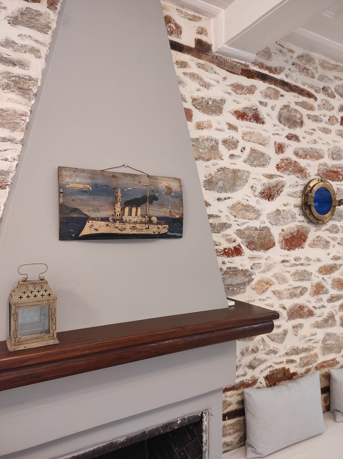 A rustic stone wall is complemented by a brown wooden mantel. A decorative ship painting is mounted above, featuring muted colors. A vintage lantern is placed on the shelf beneath the artwork, adding a classic touch. A round blue mirror enhances the decor on the opposite wall.