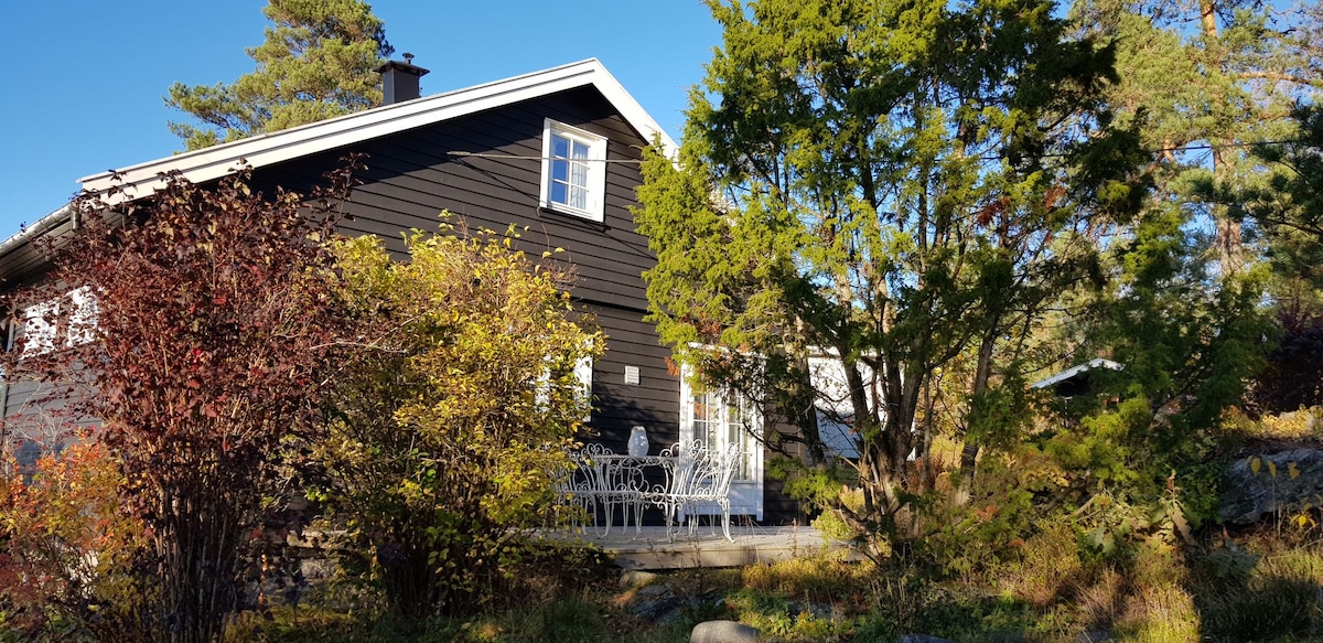 A charming wooden house is surrounded by lush greenery, featuring a spacious outdoor area with white garden furniture. Sunlight casts warm tones on the exterior, highlighting the tranquil setting that offers a blend of nature and comfort.