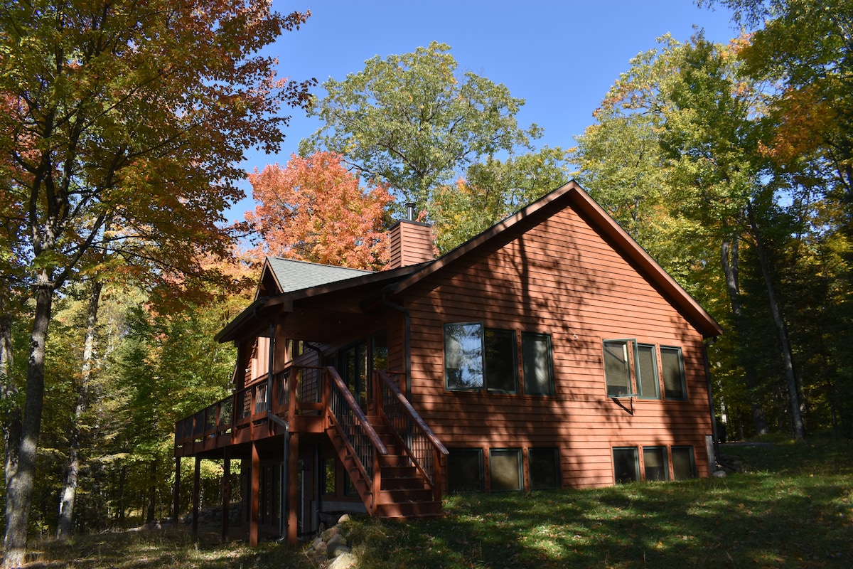 The lodge is set against a backdrop of vibrant autumn foliage, featuring a rustic wooden exterior. Large windows provide natural light, and a deck extends from the side, inviting outdoor relaxation. The surrounding trees display a mix of green and warm fall colors.