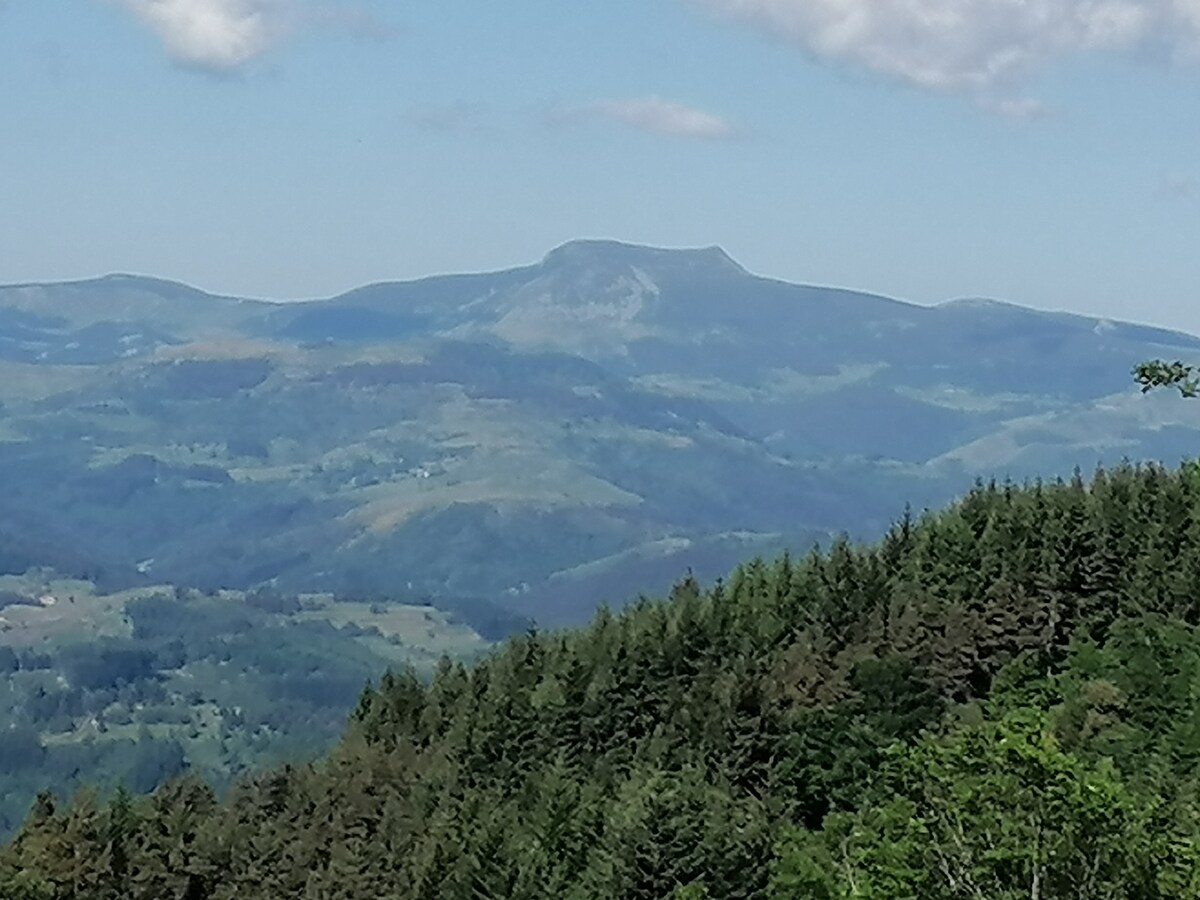 A scenic view of distant mountains is presented, with gentle slopes and patches of greenery. The landscape features rolling hills in varying shades of blue and green, showcasing the natural beauty of the countryside under a clear sky with a few scattered clouds.