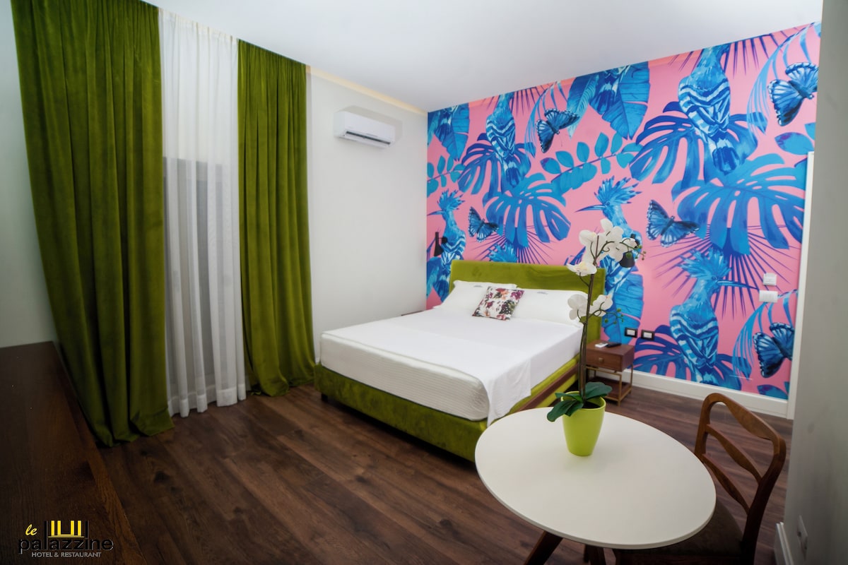 A modern bedroom features a spacious bed with a green upholstered headboard. A vibrant pink wall showcases tropical leaf patterns, while a small table sits next to the bed. Natural light filters in through a window draped with sheer curtains.