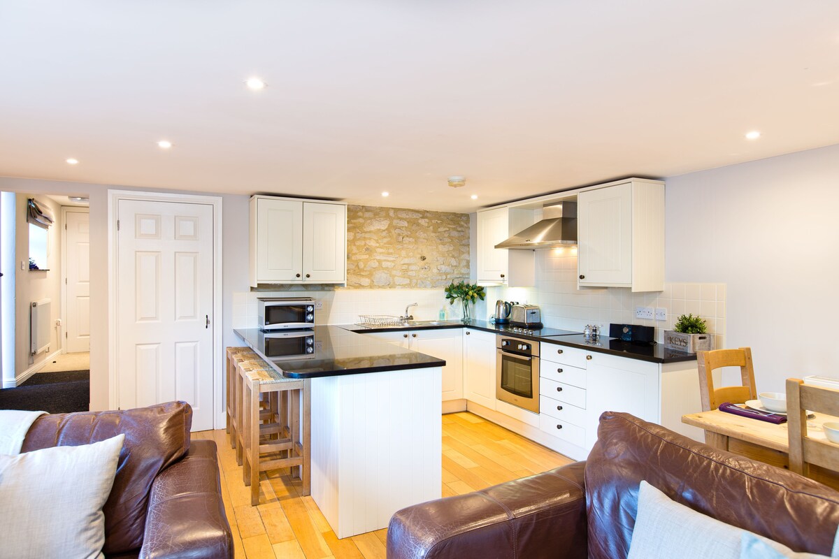 An open-plan kitchen and living area features modern cabinetry and a contrasting dark countertop. Stainless steel appliances are neatly arranged, while a wooden dining table and chairs provide seating. Natural wood flooring adds warmth, complemented by soft leather sofas positioned for relaxation.