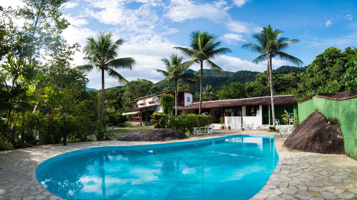 A large pool invites relaxation, surrounded by lush greenery and palm trees. A two-story building is visible in the background, with an outdoor patio area furnished with chairs and tables. The landscape features rolling hills and a serene sky, completing a tranquil setting.