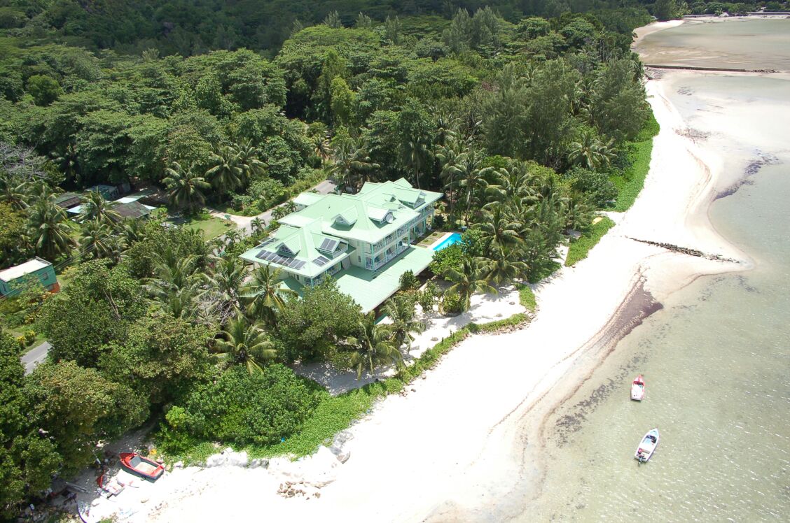 An aerial view captures a spacious beachfront property surrounded by lush greenery and palm trees. The white sandy beach gently curves along the coastline, with boats visible in the clear water. The building features a green roof, blending harmoniously with the natural environment.