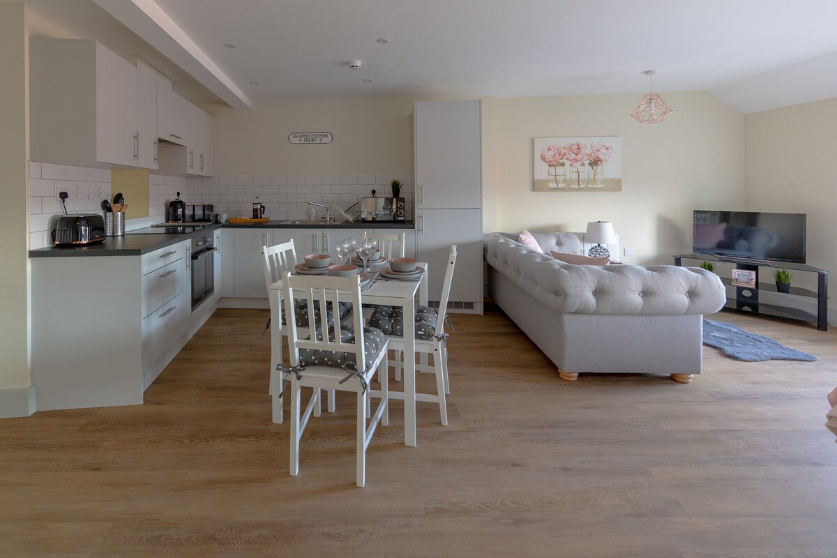 An open-plan living area features a dining table with seating for four and a modern kitchen equipped with integrated appliances. A spacious sofa is positioned nearby, facing a flat-screen smart TV, with natural light illuminating the warm wood flooring.