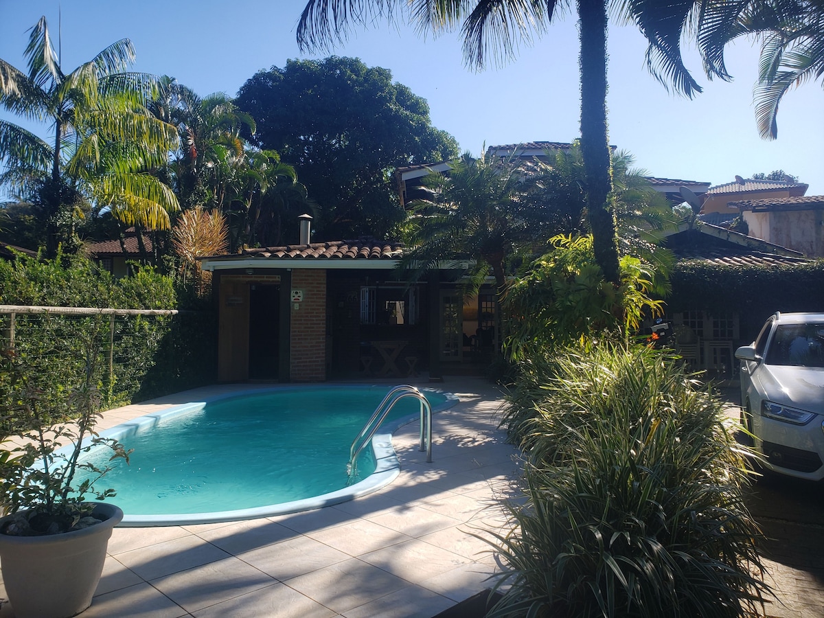 A private pool area is seen surrounded by tropical greenery and palm trees. The pool features a gentle curve and steps leading into the water. A parking space for a vehicle is visible nearby, and the house is partially obscured by lush foliage.