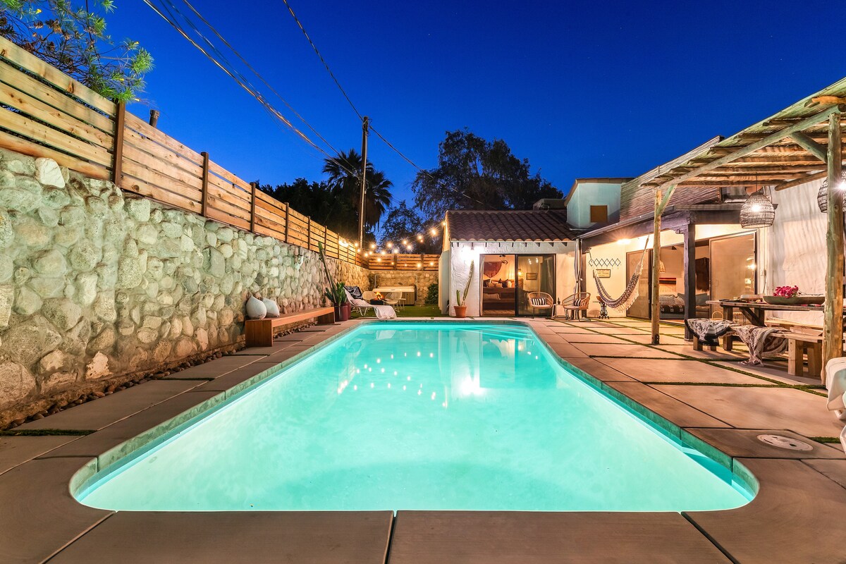 A serene pool area is illuminated by soft lights, reflecting off the clear water. Stone walls frame the space, while a cozy seating area under a pergola invites relaxation. Nearby, large glass doors seamlessly connect the inside to the outdoor retreat.