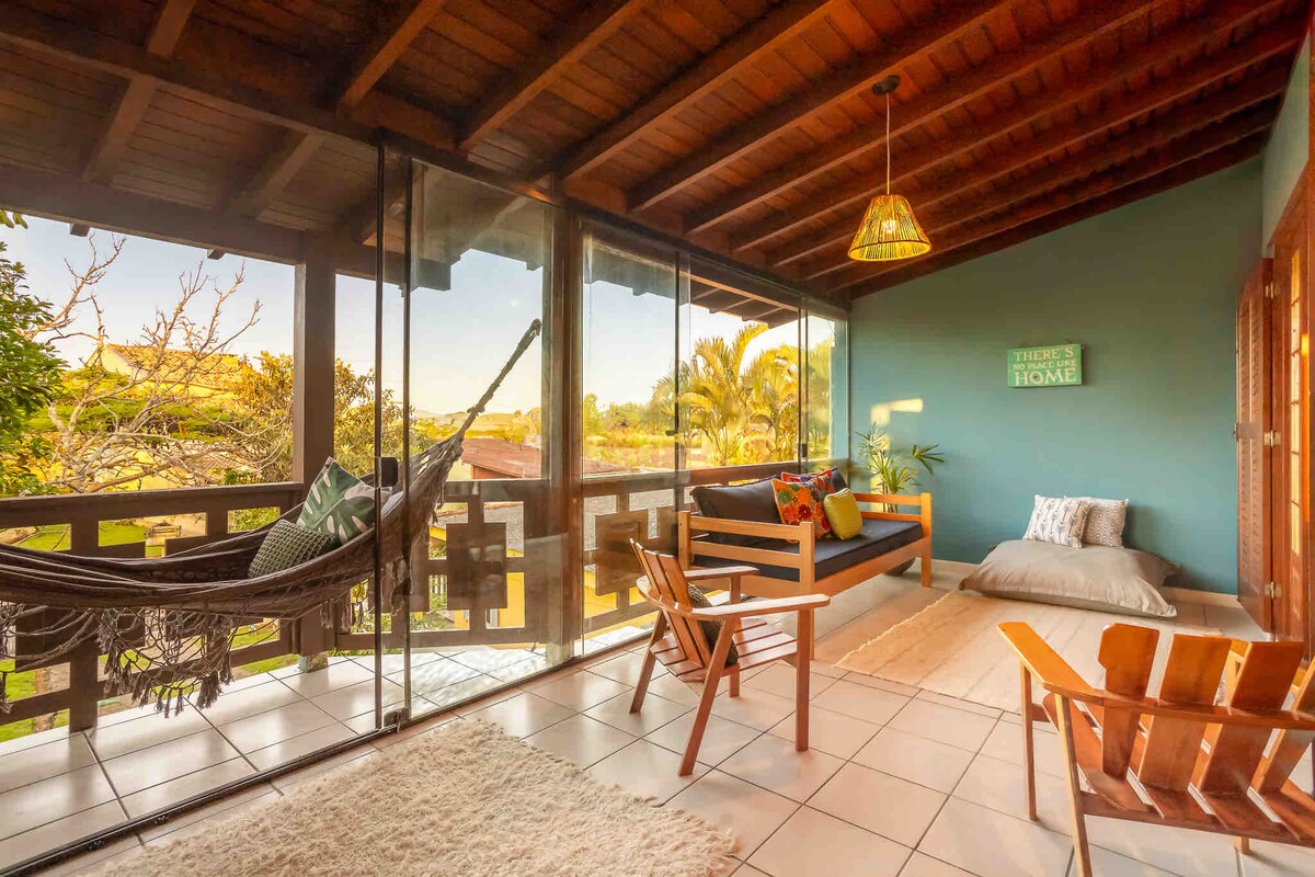 A sunny sitting area features a comfortable hammock and a wooden chair positioned beside a large window, offering views of greenery. Natural light fills the space, which is adorned with a cozy rug and a decorative wall sign. A simple cushion rests on the floor.