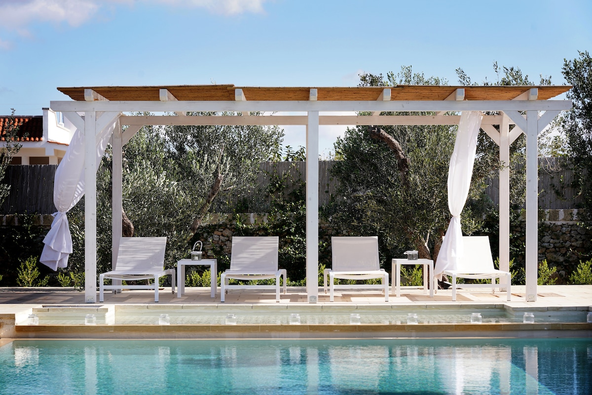 A covered outdoor seating area is positioned beside a serene pool, featuring four white lounge chairs and a small table. Light, flowing curtains are softly draped, and surrounding olive trees lend a touch of greenery to the peaceful setting.
