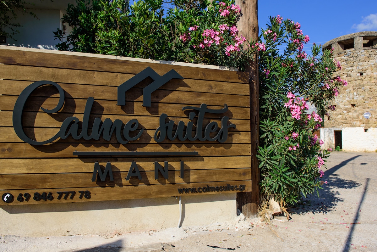 A wooden sign displays the name 'Calme Suites Mani', surrounded by greenery and blooming flowers. The rustic design blends with the stone walls of the nearby building, suggesting a welcoming atmosphere. The blue sky above enhances the vibrant colors of the scene.