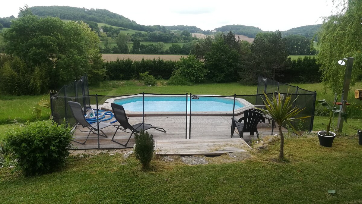 A serene swimming pool is surrounded by a wooden deck and a safety fence. Lounge chairs are positioned on the deck, facing the water. Lush greenery and rolling hills are visible in the background, enhancing the tranquil outdoor setting.