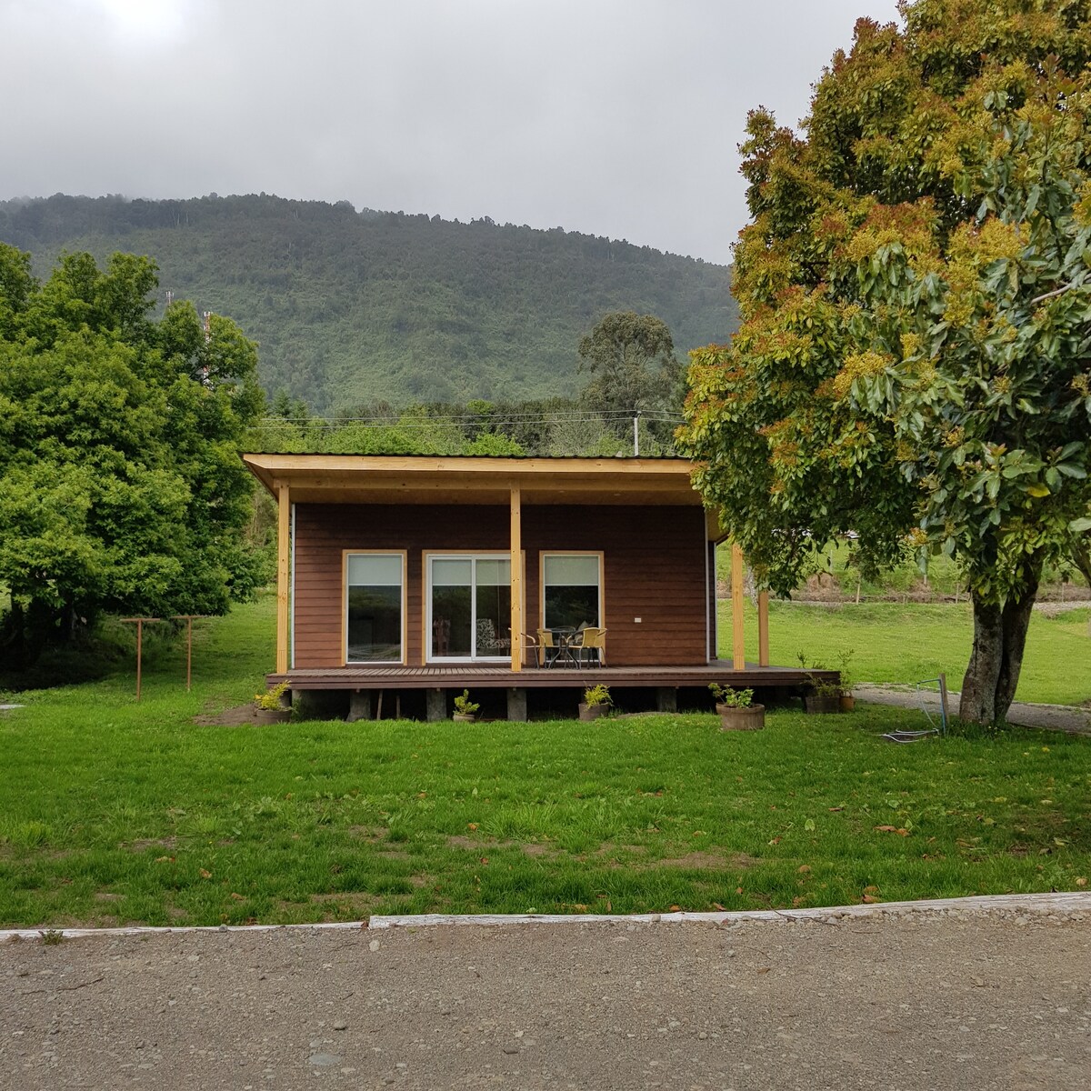 The cabin, built with wooden materials, is set on a grassy area surrounded by trees. Large glass doors and windows provide visibility of the interior. A covered porch extends outward, inviting relaxation while enjoying the natural landscape of the nearby mountains.