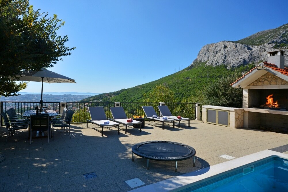 An outdoor space features a spacious patio with sun loungers and a dining set under a shaded umbrella. A trampoline is included, alongside a barbecue area with a stone grill. The scenic view encompasses rolling hills and the distant landscape.