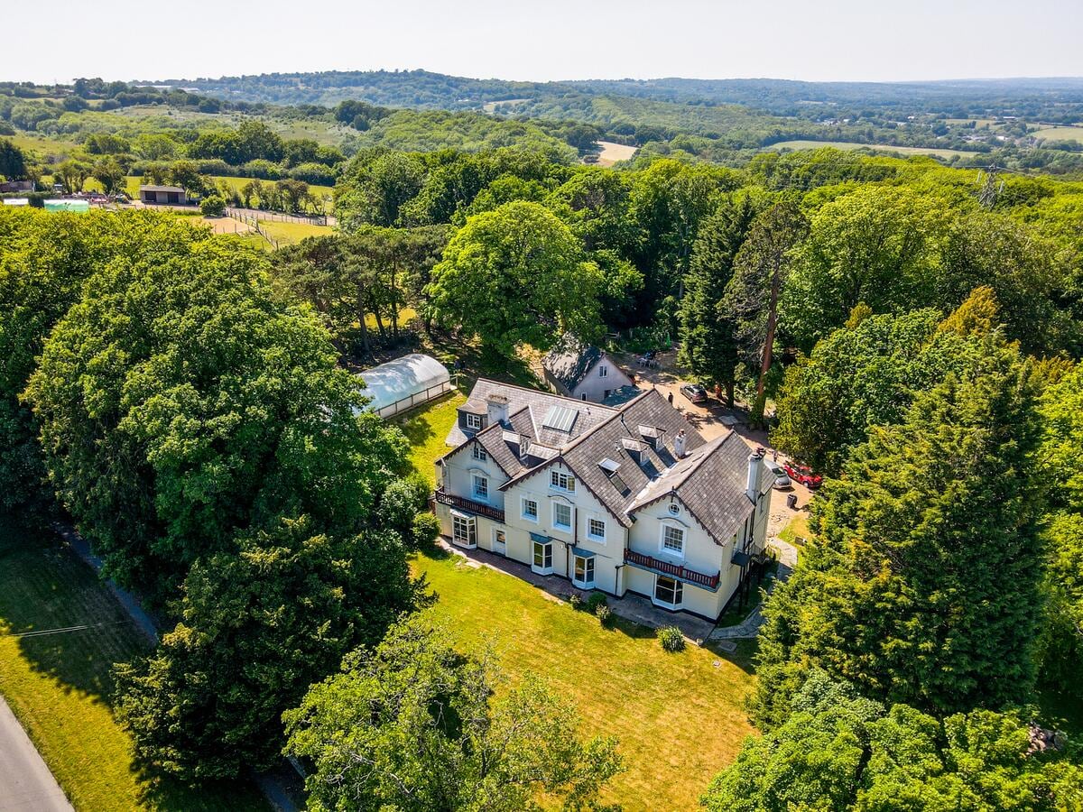The spacious property is nestled among lush greenery, surrounded by trees and expansive lawns. A harmonious blend of architecture is visible, showcasing multiple levels and a varied roofline under clear blue skies. The tranquil natural landscape offers a serene backdrop.