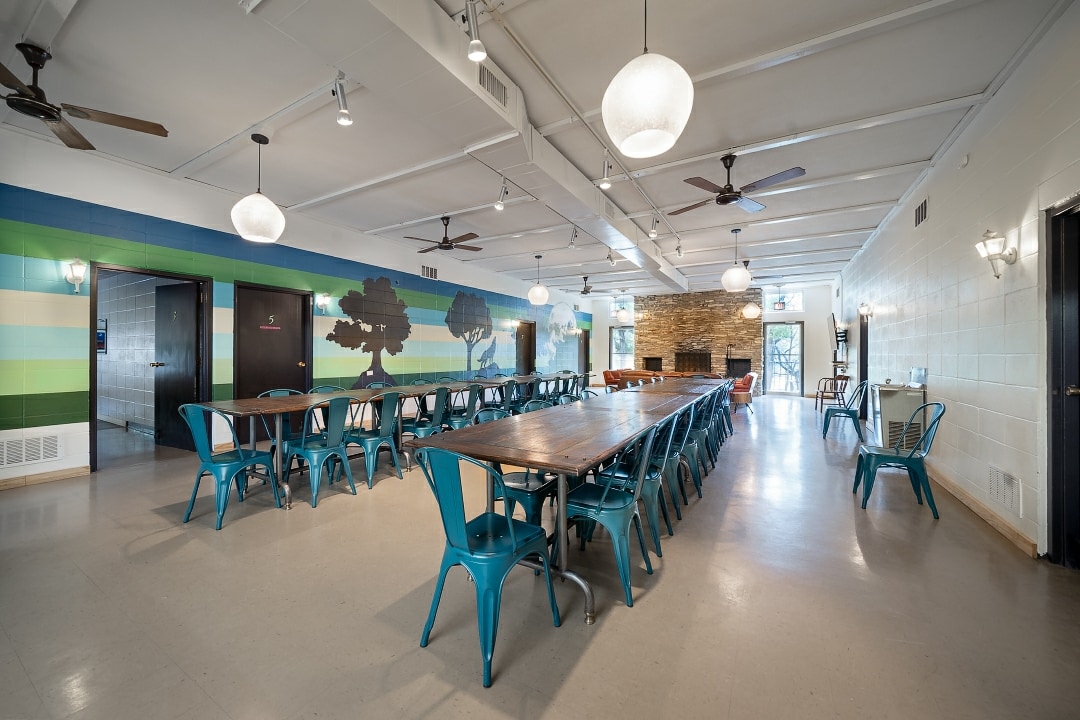 A spacious dining area is furnished with long tables accompanied by teal chairs. Colorful murals line the walls, depicting trees and nature. Two ceiling fans and multiple light fixtures ensure a well-lit environment, enhancing the room's functional design for group gatherings.
