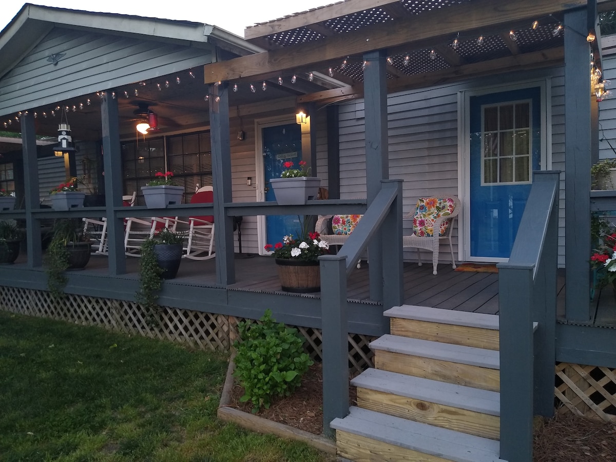 A charming porch features two rocking chairs and colorful flower pots, creating an inviting space. The entrance is marked by a bright blue door. String lights are hanging above, adding a warm touch to the evening ambiance. Steps lead to the grassy yard.