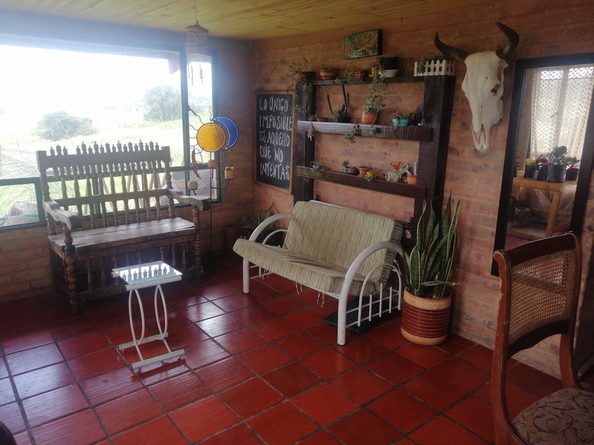 A cozy indoor sitting area features rustic wooden furniture, including a rocking chair and a white loveseat. A wall shelf adorned with plants and decorative items adds character. Large windows invite natural light and provide views of the surrounding landscape.
