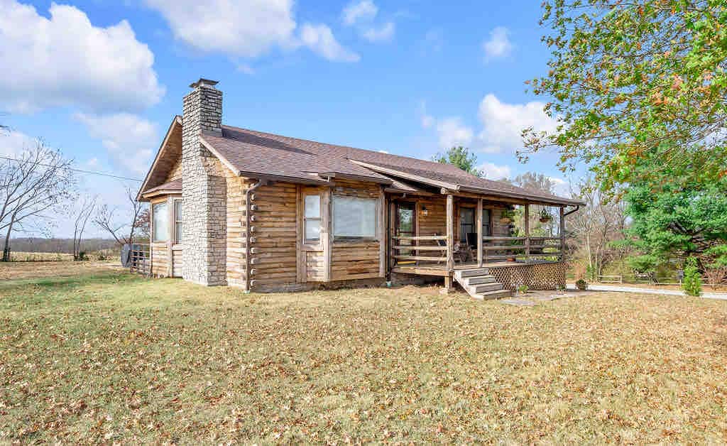 A charming log cabin is set against a clear blue sky, featuring a stone chimney and a welcoming front porch. The surrounding landscape showcases a grassy yard dotted with fallen leaves, and trees are visible in the background, enhancing the cabin's rustic appeal and connection to nature.